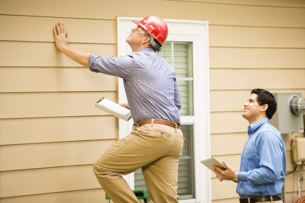 Repairmen, building inspectors, exterminators, engineers, insurance adjusters, or other blue collar workers examine a building/home's exterior wall and foundation. One wears a red hard hat and clear safety glasses and holds a clipboard. The other holds a digital tablet.